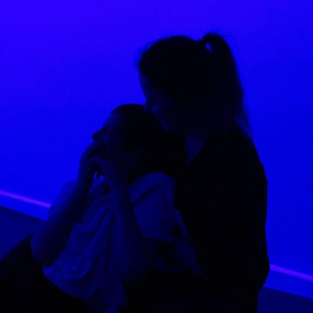 Treloar's student being held by his assistant - both sitting on a mat on the floor during an atmospherics session. The room is lit by a blue light. The student looks mesmerised by this sensory experience.