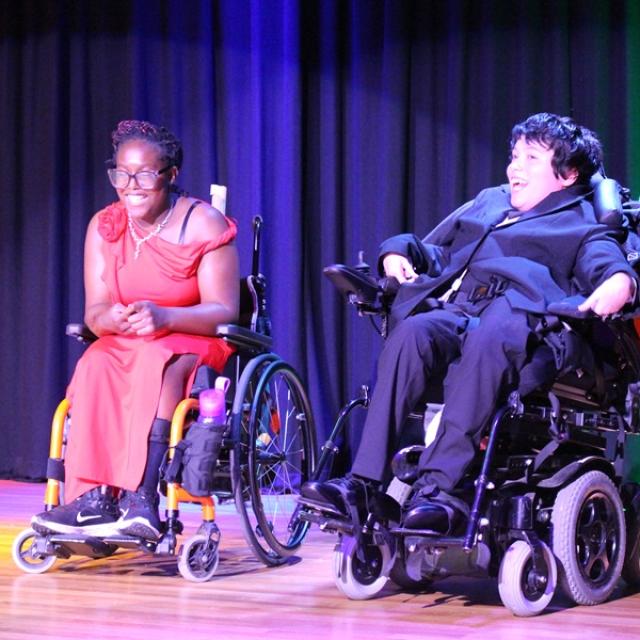 2 Treloar College students on the stage during Treloar's Got Talent competition. The stage is lit up with colourful lights. The students have just finished their performance and are looking at the audience, smiling. They are both smartly dressed: one is in a red dress, the other in a black suit.