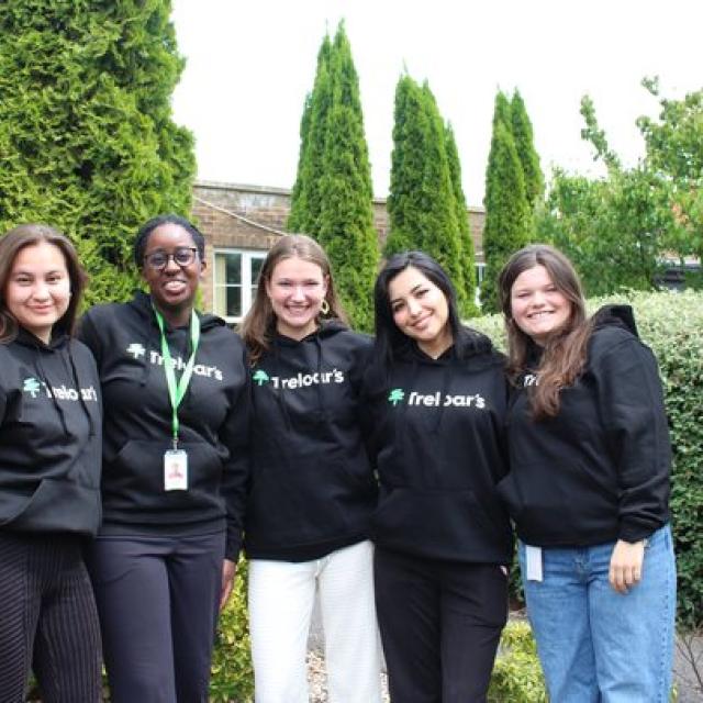 Five female volunteers standing outdoors in front of tall green shrubs, all wearing black hoodies with the 'Treloar's' logo on them.