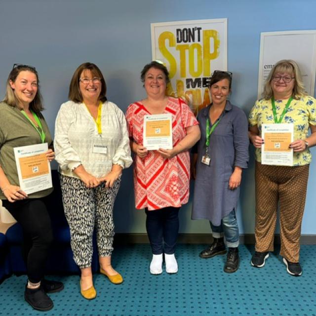 A group of 5 women, lined up in a room, posing for a picture. Some of them are holding certificates. Everyone is smiling.