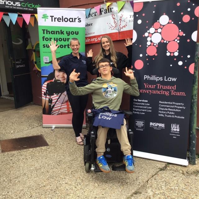 Treloar's student at the Dummer Fair posing for a photo with a sponsor and Treloar's member of staff in front of 2 banners. Everyone is waving at the camera.