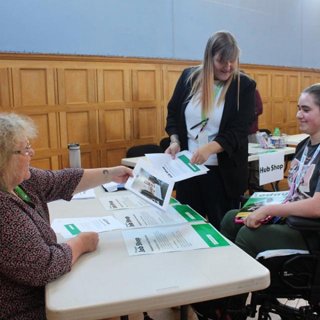2 Treloar's staff members engage in a discussion with Treloar's student at a table covered with informational brochures inside a wood-paneled room.
