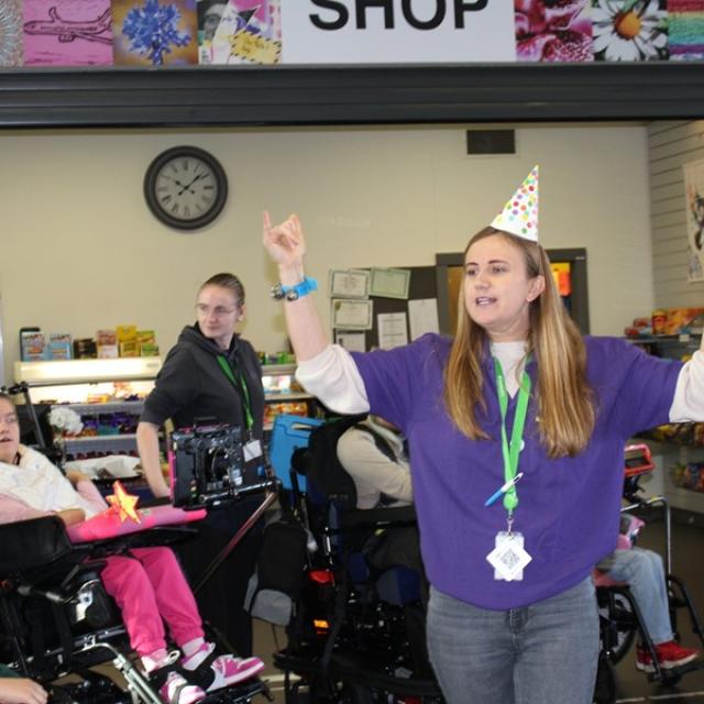 Treloar's Makaton tutor wearing a party hat and purple shirt joyfully raising her arms during a Makaton celebration event with with staff and students around her.