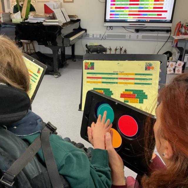 Over the shoulder shot of Treloar's student using a tablet with 3d printed case on which allows the student to play music using Clarion; the student is touching colored buttons on the screen to create music, she is guided by her assistant in a music classroom with a piano and sheet music on stands.