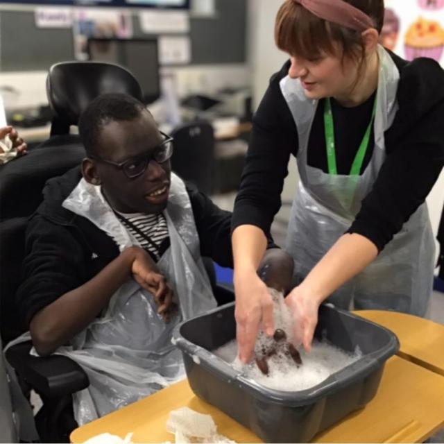 Treloar College student during his session with his occupational therapist: they are in the classroom and the OT is helping him wash his hand in a bowl with water with soap.