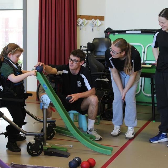 Treloar's primary student in the standing frame playing boccia with college students from a local college assisting her and observing her use a ramp.