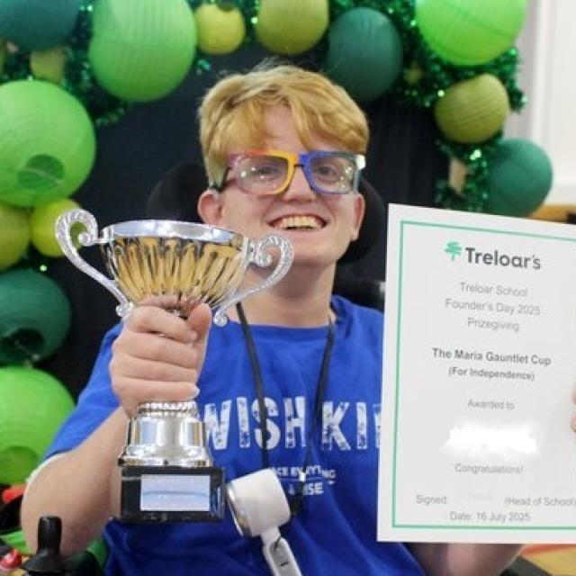 School student holding certificate and trophy under a green balloon arch