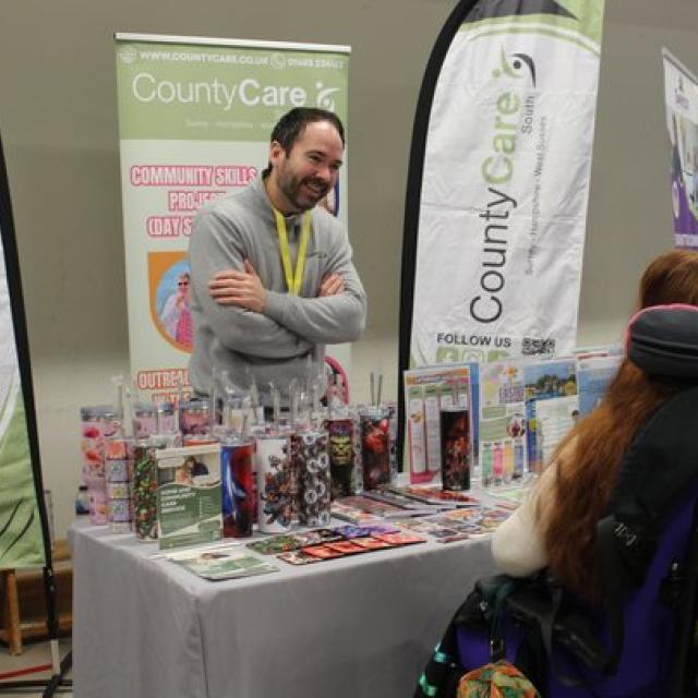 An exhibitor standing behind a CountyCare information booth with brochures and promotional items displayed on the table. He is smiling and listening to Treloar's student in front of him.