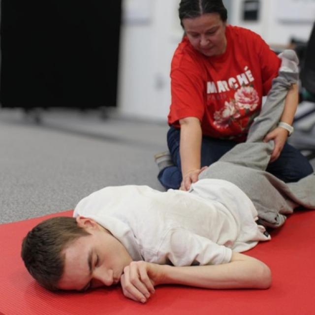 Treloar College student lying face down on a red mat while a staff member in a red shirt gently holds their leg, during a therapy / exercise session.