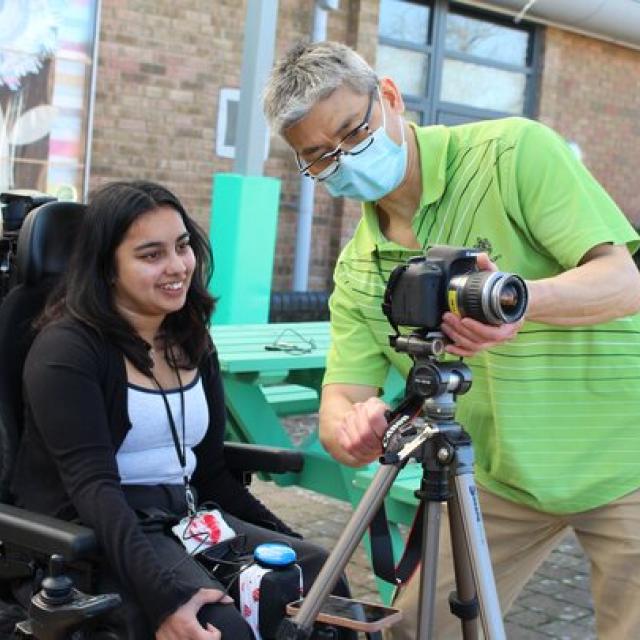 Treloar's student support assistant in green shirt is adjusting a camera on a tripod outdoors for Treloar College student who is about to take some pictures.