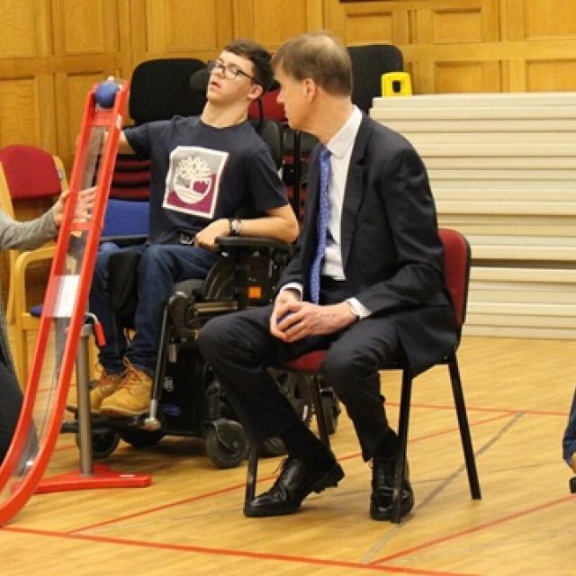 A support assistant kneels beside Treloar's student is demonstrating how to play boccia to Sir Stephen Timms who is wearing a suit and is watching attentively the student.
