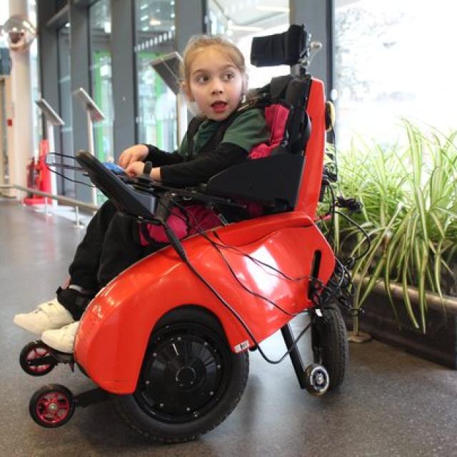 Treloar's primary student seated in a tiny trax chair wheelchair inside a spacious building with large windows and plants nearby.