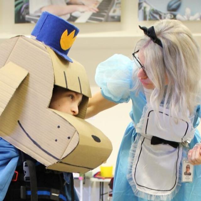 Treloar's primary student using a walker, wearing a large cardboard dog head and a hat, interacting with his Student Support Assistant dressed as Alice from Alice in Wonderland in a gymnasium.