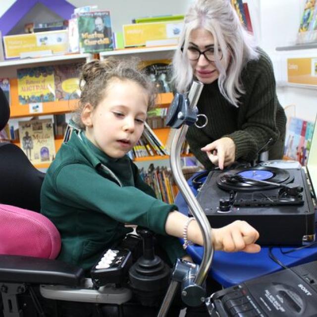 Treloar's primary student interacting with a record player while her assistant guides her in a colourful library setting.