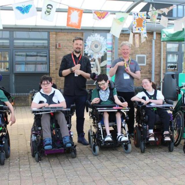 Group of 7 individuals, 5 Treloar's students and  2 adults outdoors under colourful hanging artwork in a courtyard.