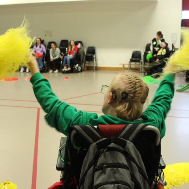 Treloar's student with braided hair enthusiastically waving yellow pom-poms in a gymnasium filled with seated spectators. He is wearing a cochlear implant.