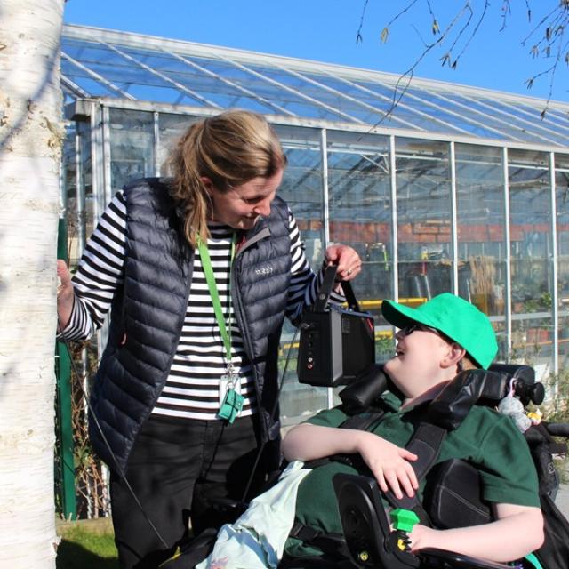 Treloar's teacher in a striped shirt and black vest smiling and interacting with a student outside a large greenhouse on a sunny day. She is holding a speaker near the boys ear so that he can listen to the tree and the sounds of transpiration.