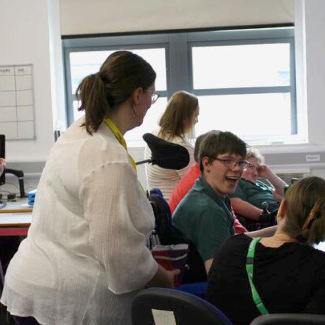 Two visitors with  yellow lanyards standing next to Treloar's students who are working on their computers in a classroom. One of the students is smiling and having a lively conversation with the visitor and his assistant.