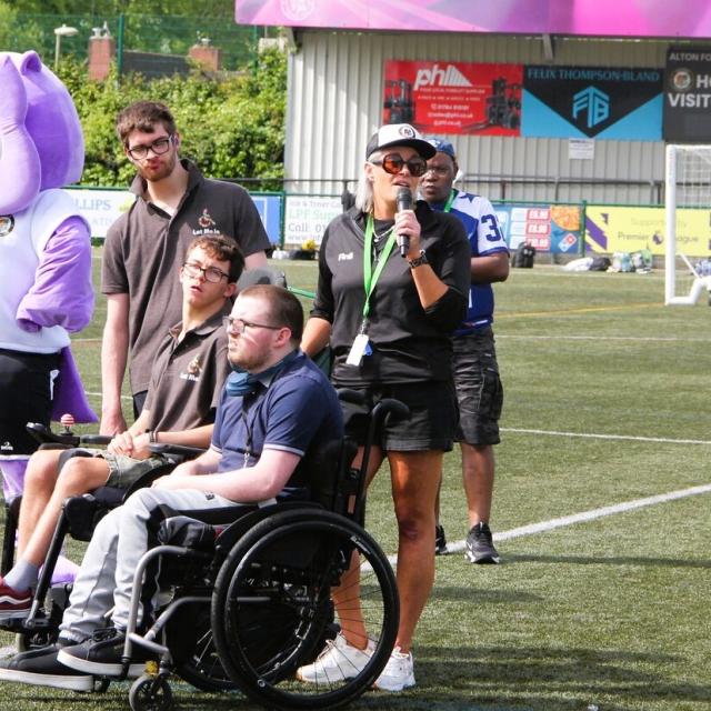 Group of people on a football field, including a Treloar's students and staff and a mascot in a purple costume, with one woman speaking into a microphone.