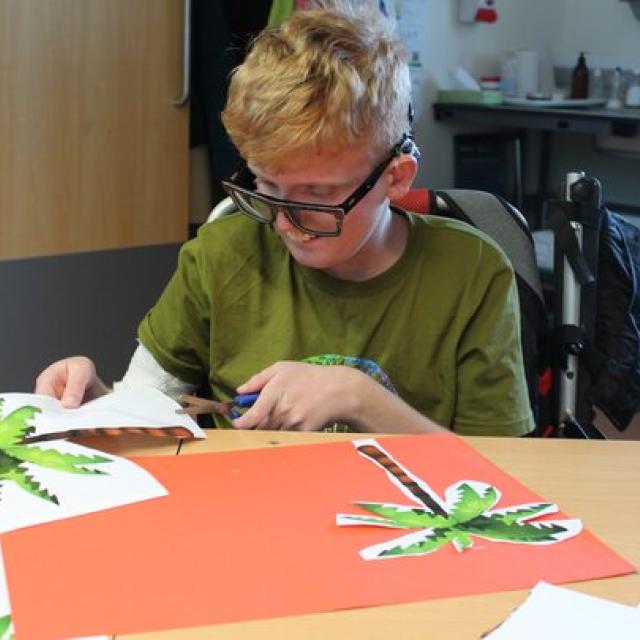 Treloar's student in a green shirt sitting at a table cutting out paper palm tree shapes for a craft project.