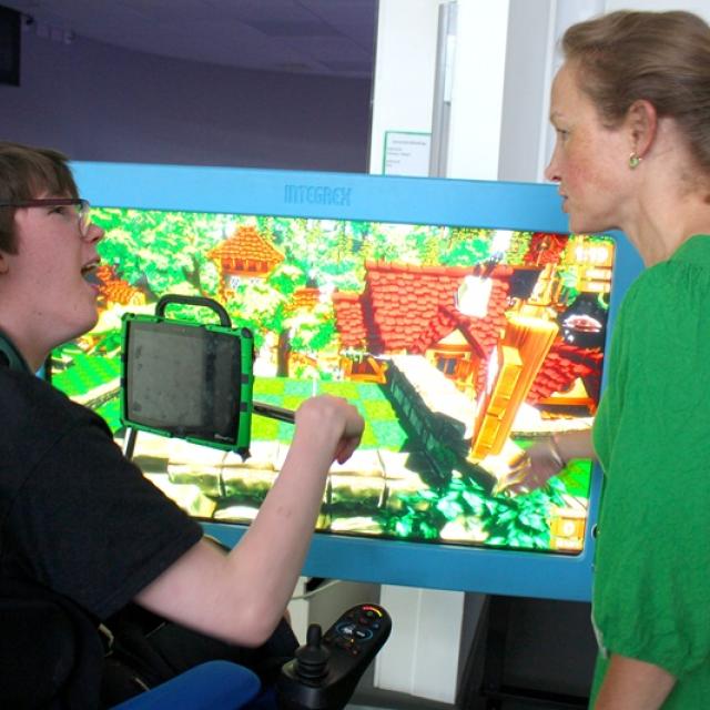 Treloar's student in front of a big screen displaying a colourful video game with a teacher in a green dress having a discussion about the game.