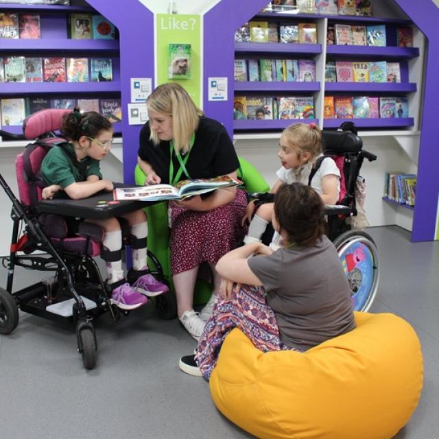 Teacher reading a book to two Treloar's primary students and a support assistant sitting on a yellow beanbag in a colourful library.