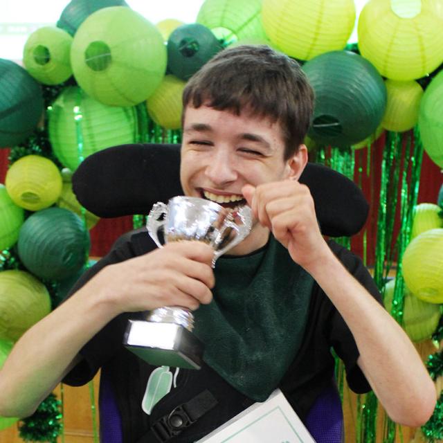 Smiling Treloar's student holding a silver trophy with green paper lanterns in the background. He is very happy and proud of the award.