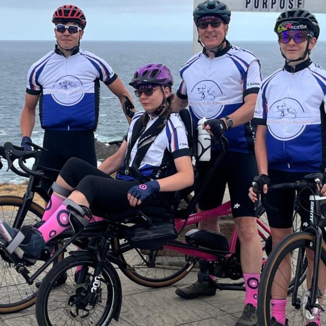 A family of four cyclists stand together in front of a sign post.