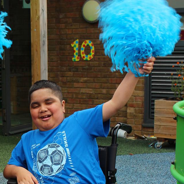Smiling Treloar's primary student waves a large blue pom-pom outdoors near a colourful number 10 on a brick wall.