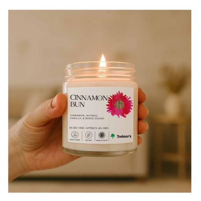 Hand holding a lit candle labeled Cinnamon Bun with scents of cinnamon, nutmeg, vanilla, and baked dough.