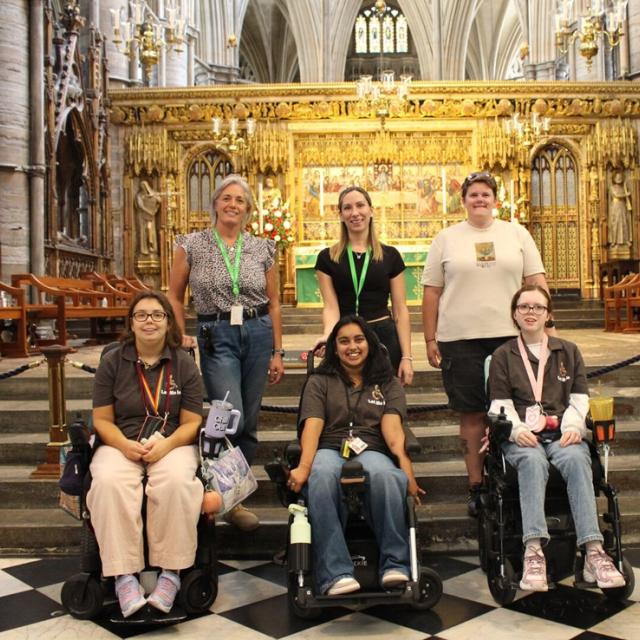 Group of six people, including three in wheelchairs, posing inside a grand cathedral with ornate golden altar in the background.