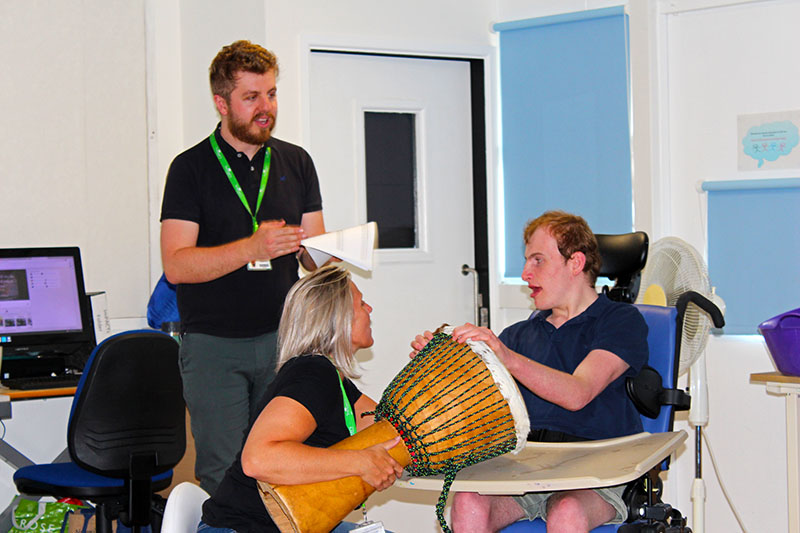 Teachers with a student during a sensory activity: teacher holding a drum and student is tapping the drum with his hands