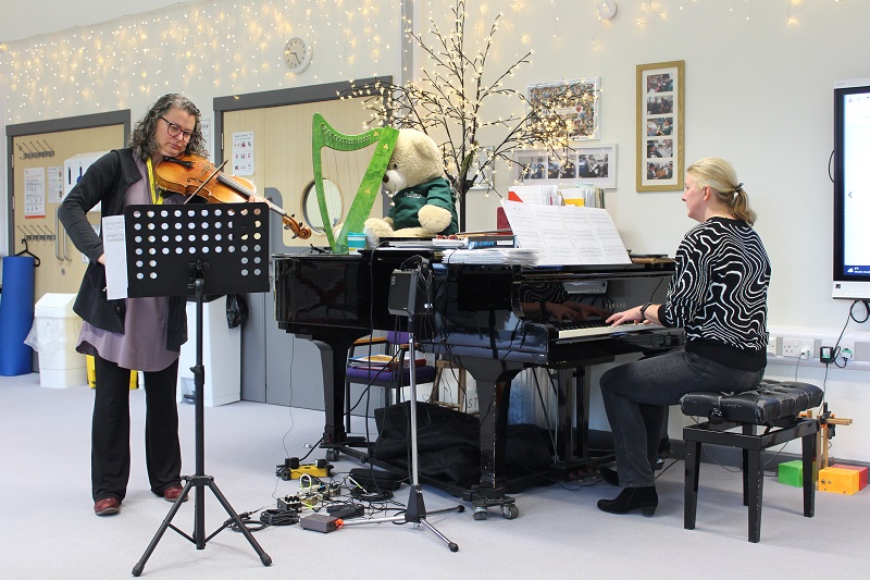 Viola workshops at Treloar School: Kate Read, a professional viola player, performing for students in the classroom full of fairy lights, standing by the piano. Treloar's head of Music is playing the piano.