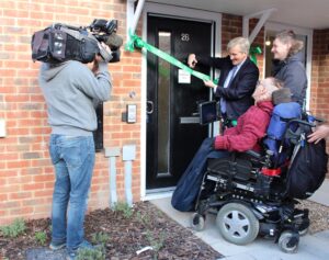 East Hampshire MP, Rt Hon Damian Hinds cutting the ribbon to the new flat while being filmed by a camera man; new tenants watching Damian cut the ribbon