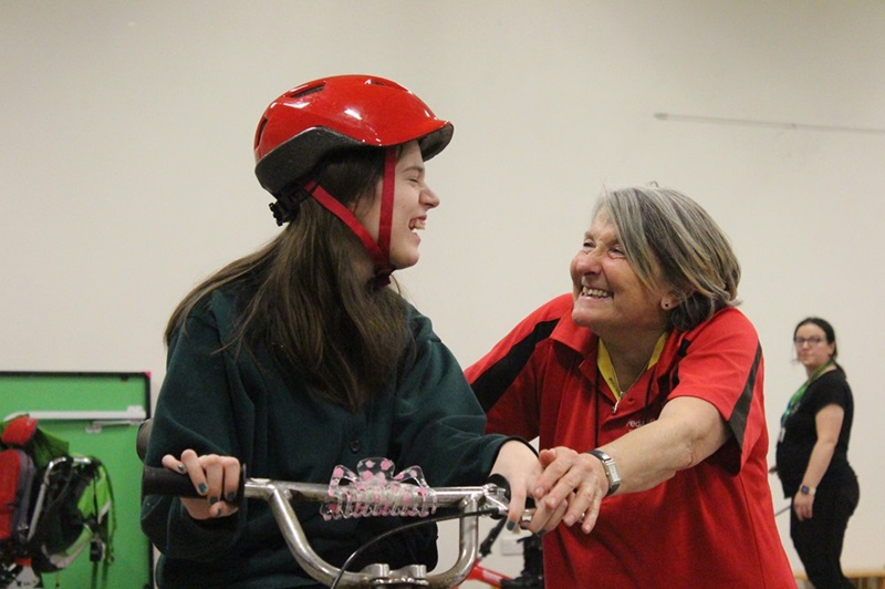 Treloar's student while triking, holding her both hands on the handlebars, wearing shiny red helmet, looking at the instructor who is helping her control the trike. Both smiling at each other.